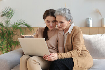 Happy elderly middle mother sitting on couch with her daughter, looking at laptop. Young woman showing video, photos to mommy, trusted relations. Family concept.