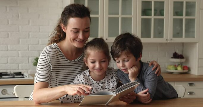 Smiling young beautiful mother or female babysitter reading fairy tale or interesting story from paper book to joyful adorable little children siblings in kitchen, watching colorful pictures together.