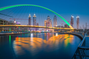 Fototapeta premium Illuminated in green color Tolerance Bridge in Dubai with an unusual architectural design against the backdrop of skyscrapers. Urbanism and sightseeing in the UAE