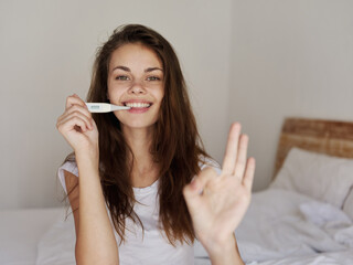 joyful woman sitting out of bed with a thermometer in her mouth positive result