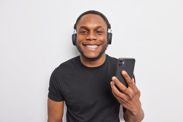 Cheerful dark skinned man smiles pleasantly enjoys listening music from playlist holds modern smartphone uses wireless headphones wears black t shirt isolated over white background. Technology