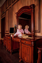 Obraz na płótnie Canvas Pretty one-year-old girl in pink dress with an old book is sitting on sideboard by mirror. Child in library interior. Concept of education and child rearing. Background for site or banner. Copy space