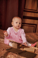 Pretty one-year-old girl in pink dress with an old book is sitting on bed in room. Child in bedroom interior. Concept of education and child rearing. Background for site or banner. Copy space