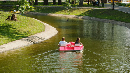 Two girls ride a red catamaran on a pond in the park. Selective focus.