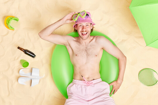 Relaxed topless bearded man laughs happily wears panama and shorts snorkeling mask enjoys subathing spending summer vacation at beach looks positively surrounded by slippers beer green parasol