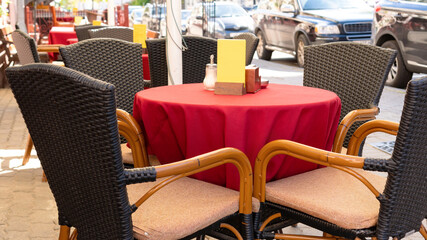 Table with red tablecloth and rattan chairs in a street city cafe on a summer day. Cozy table in a cafe close-up with a menu with copy space and a salt shaker.