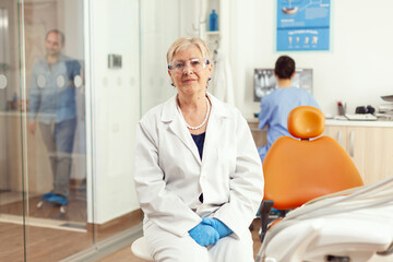 Obraz premium Orthodontist senior in medical uniform sitting on chair looking into camera waiting for man pacient to start stomatology treatment after tooth surgery. Team of dentists working in cabinet office