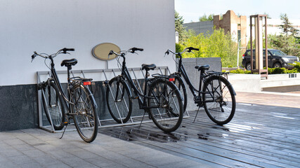 Fototapeta na wymiar Bicycle rental service in the hotel. Classic black bicycles are parked in the metal bike parking in the hotel lobby on a wooden deck. Bicycles stand in a puddle after a summer rain.