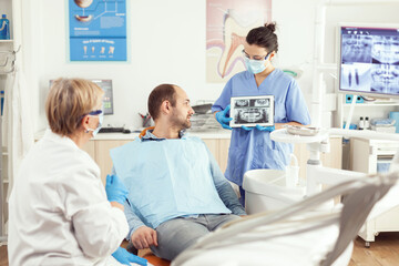 Obraz premium Sick man sitting on stomatological chair listening doctor while looking at tablet in dental clinic. Dentistry doctor showing to patient teeth radiography during dental consultation