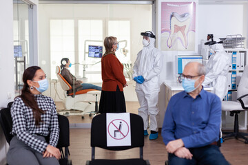 Naklejka premium Senior woman patient in stomatology clinic discussing with doctor dressed in ppe suit as safety precaution during global pandemic with coronavirus. People talking in stomatology waiting room.