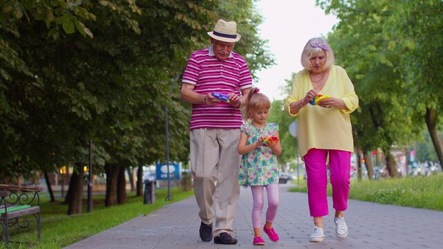 Senior grandmother grandfather walking with granddaughter playing squeezing anti-stress silicone toy simple dimple game in park. Family with pop it bubble squishy game with buttons. Stress reliever