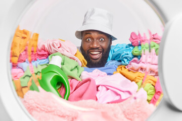 Cheerful dark skinned husband wears panama on head poses from inside of washing maching feels positive does laundry has busy day overloaded with multicolored dirty clothes. Household routine
