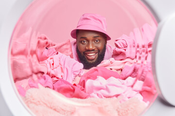 Bearded cheerful African American man photographed from inside of washing machine loads washer with dirty laundry wears panama. Everything in pink color. Home lifestyle and laundering concept