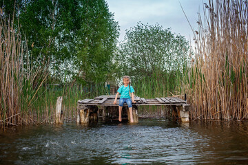 Cute preteen girl sits on a wooden pier by the lake and stirs feet in the water, happy childhood...