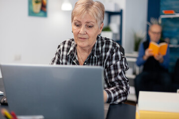 Senior lady entrepreneur working and pointing at computer screen while browsing on internet. Elderly woman in home living room using moder technoloy laptop for communication sitting at desk indoors.