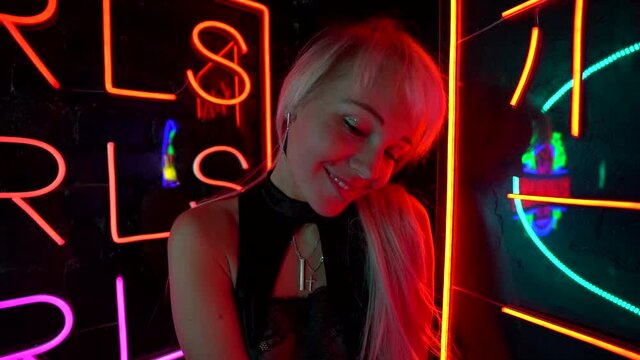 a blonde in a black top and denim shorts is standing in a dark room with neon signs. she smiles and touches her long hair. the camera is moving