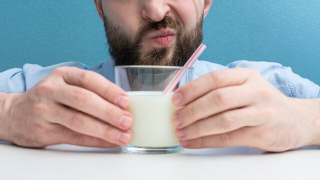 Man doubts whether he needs to drink cow's milk, bearded with glass of milk with a straw, cropped image, close-up, 16:9