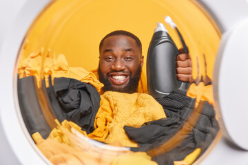 Positive dark skinned man photographed from inside of washing machine holds bottle of liquid detergent surrounded by piles of clothes poses in laundry room. Busy housekeeper does domestic chores