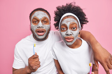 Surprised Afro American couple look with wondered expressions at camera wear clay beauty masks on face dressed in casual t shirts hold toothbrush isolated over pink background. Skin care concept
