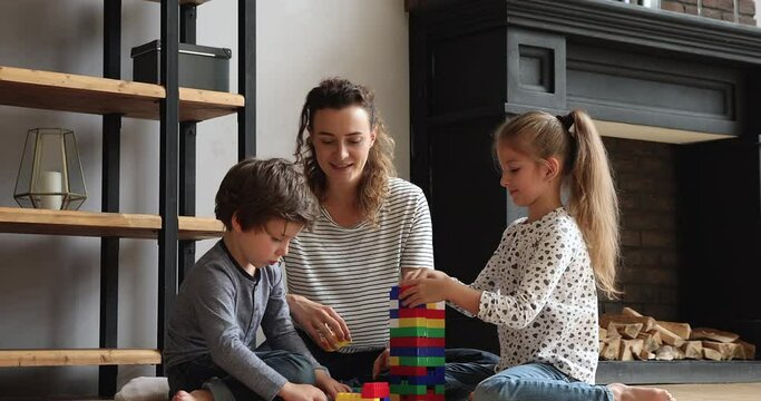 Smiling beautiful young mother on babysitter playing with joyful small children boy and girl in modern living room, constructing building with colorful plastic blocks, daycare entertainment concept.
