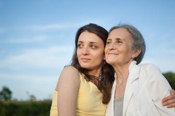 Obraz premium Senior mother with gray hair with her adult daughter looking at the camera in the garden and hugging each other during sunny day outdoors