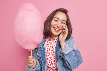 Positive carefree pretty Asian teenage girl smiles sincerely enjoys spare time eats sweet cotton...