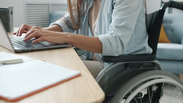 Unrecognizable disabled girl is using laptop typing sitting in wheelchair at desk busy with online work indoors at home. Technology and lifestyle concept.