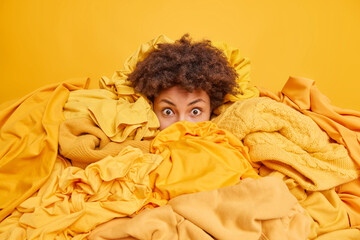 Scared curly haired Afro American woman covered with pile of clothes cleans out wardrobe tries to...