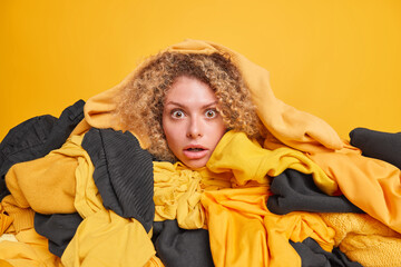 Emotional woman with curly hair chooses what to wear poses among pile of clothes stares impressed...