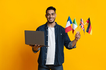 Cheerful arab guy holding laptop and international flags on yellow