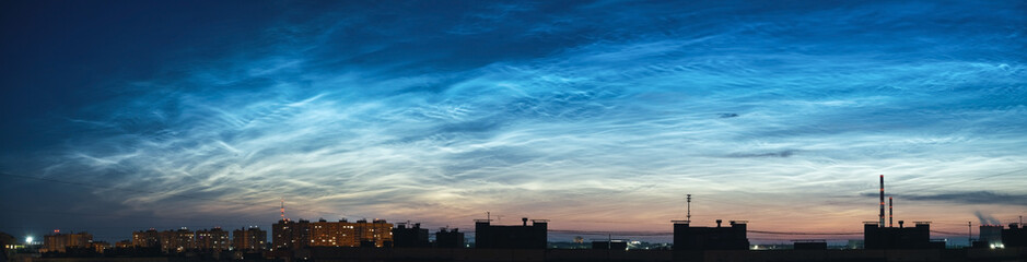 Panorama of the night sky and spectacular silvery clouds, silhouettes of city buildings against a background of shining clouds. Atmospheric phenomenon glowing of noctilucent clouds.