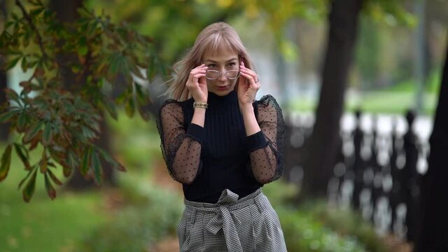 a blonde in an elegant black blouse and plaid trousers puts on glasses and laughs under a chestnut tree against the background of a blurred openwork lattice of the park