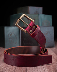 A red leather belt with a bronze buckle rests on a wooden table against a leather goods background