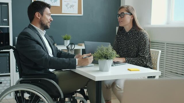 Disabled guy in wheelchair is discussing business with female colleague talking sitting at desk in office. Corporate communication and disability concept.