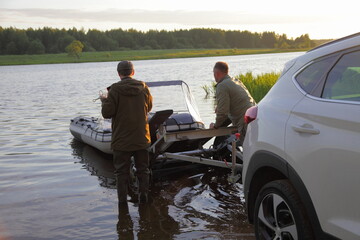 Two European man unloads an inflatable motor boat from a boat trailer into the water before fishing recreation, resting on the Russian river at summer morning