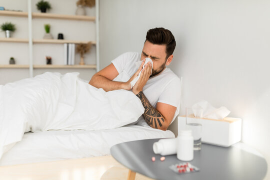 Young ill man blowing runny nose in paper tissue, sneezing, having cold symptoms, lying in bed at home interior