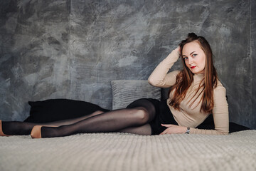 Naklejka premium A beautiful young woman with red lipstick and brown hair sits sideways on a large bed against the background of a dark wall. Stretched out her legs in black tights and beige shoes.