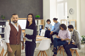 Portrait of confident female and male business partners standing in office against workers background. Young woman and middle-aged man smiling and posing in front of the camera before negotiations.