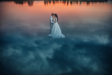 Newlywed couple standing on water at sunset.
