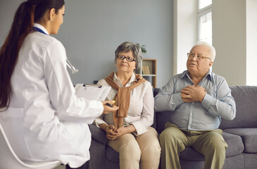 Mature couple sitting on a sofa talking and consulting about health with a doctor during her visit. Female doctor listens to the elderly and records the information in a medical record.