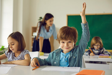 Portrait of smart clever primary school student sitting at desk in classroom raising hand smiling to camera. Pick me gesture. Positive children emotion. Back to school and education concept - Powered by Adobe