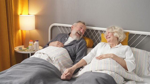 Positive caucasian elderly couple have fun. Laugh while lying on bed at home, in domestic clothes, morning, daily routine of retired people, harmonious relationships.
