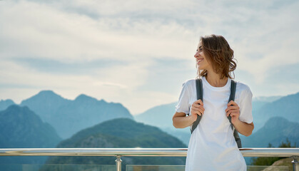 Pleased tourist female wearing white shirt with backpack standing in front of mountain top with cloudy sky, viewpoint of Tunektepe Teleferik, Antalya