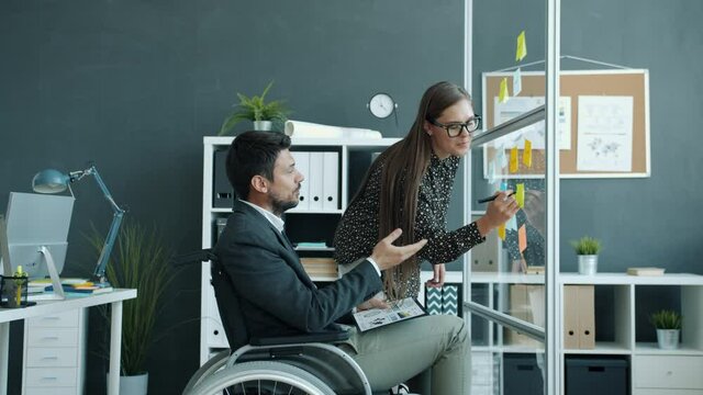 Young woman and disabled man in wheelchair are talking writing on sticky notes on glass board working together in office. Creative business and people concept.