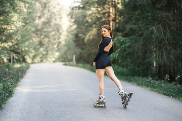 Young caucasian fit woman on roller skates posing outdoors in woods on asphalt road. Wearing open crop top and black sporty shorts. - obrazy, fototapety, plakaty