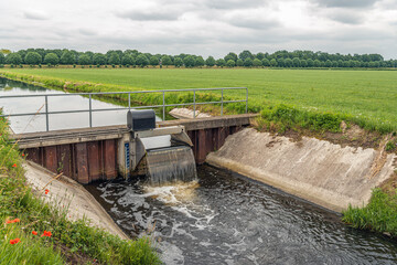 The water foams at the overflowing weir in a Dutch polder landscape. The small weir keeps the water...