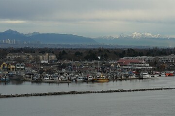Fototapeta na wymiar Fishing village of Steveston with boats on the Fraser river in , typical port town in west Canada connected with Pacific Ocean. View from container vessel and on the background are mountains.