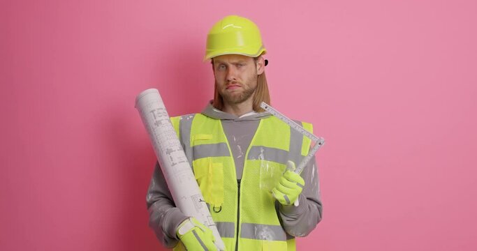 Serious attentive bearded male architect hols blueprint and tape measure looks around room does house remodeling wears uniform isolated over pink background. Engineering and maintenance concept