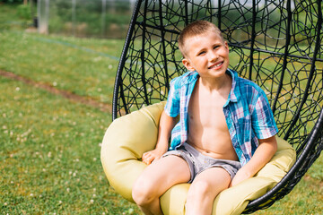 Happy boy in plaid shirt sitting in cocoon chair in the garden. Summer holidays