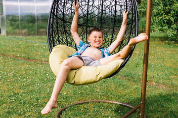 Happy boy in plaid shirt sitting in cocoon chair in the garden. Summer holidays